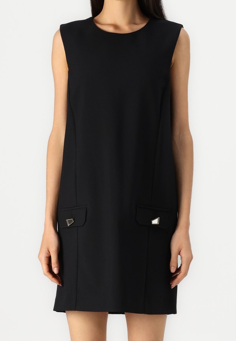 Femme portant une robe noire sans manches avec deux petites poches à rabat, debout devant un fond clair uni.