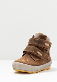 Bota de ante marrón con dos correas de velcro, puntera reforzada, entresuela gris y suela de goma con taco amarillo. Forro suave visible en la parte superior.