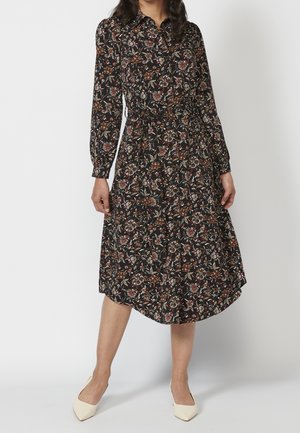 Kvinde iført en sort, brun og cremefarvet blomstret langærmet midikjole med bindebånd i taljen og cremefarvede spidse sko, stående foran en hvid baggrund.