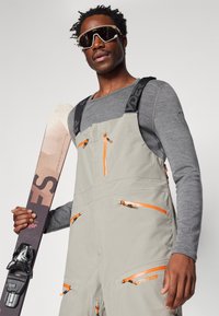 Beige skidräkt med orange detaljer och flera fickor med dragkedja, kombinerad med en grå långärmad tröja. Håller i skidor, bär solglasögon.