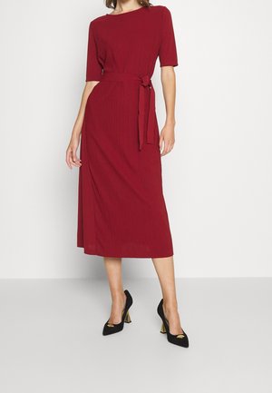 Punainen ribattu midi-mekko, jossa on lyhyet hihat, vyöllinen vyötärö, litteä kaulus ja teksturoitu kangas. Yhdistetty mustiin korkokenkiin.