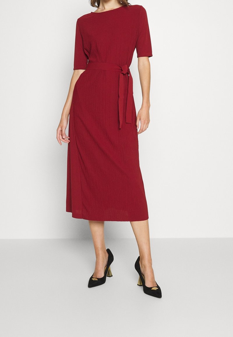 Rotes geripptes Midi-Kleid mit kurzen Ärmeln, tailliertem Gürtel, flachem Kragen und strukturiertem Stoff. Kombiniert mit schwarzen High Heels.