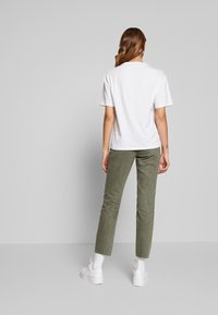 Kvinna med långt hår iförd en enkel vit t-shirt, olivgröna byxor, vita strumpor och vita sneakers som står mot en grå bakgrund.