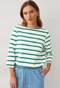 Witte langwerpige top met groene horizontale strepen, ronde hals en een relaxte pasvorm. Gecombineerd met een lichtblauwe denim rok.