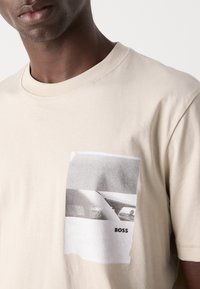 Beige bomull T-skjorte med rund hals, med en rektangulær svart-hvitt grafisk utskrift på venstre brystområde.