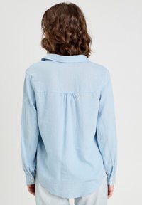 Light blue long-sleeve shirt made of soft fabric, featuring a back pleat, a classic collar, and buttoned cuffs. Smooth texture and casual fit.