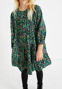 Barevné šaty se zeleným, růžovým a černým abstraktním vzorem, s dlouhými rukávy a volným střihem, nošené s černými legínami a botami.