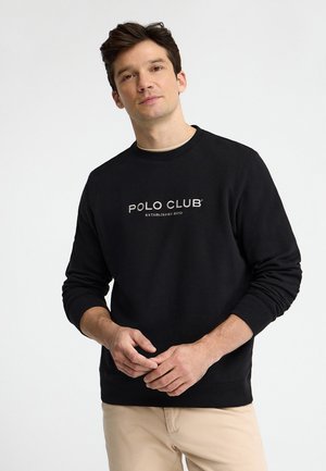 Uomo con felpa nera "Polo Club" e pantaloni beige, in piedi con le mani leggermente giunte davanti a uno sfondo bianco.
