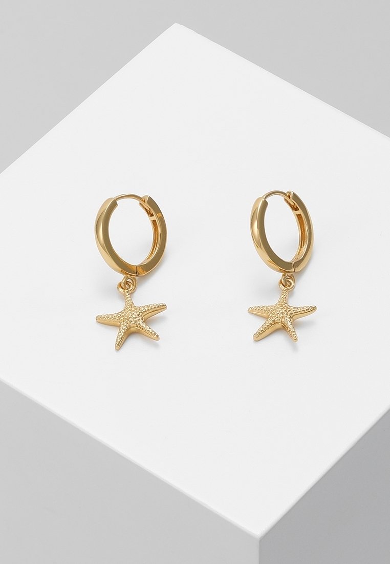 Boucles d'oreilles créoles en or avec des breloques texturées en forme d'étoile de mer, suspendues, exposées sur une surface blanche inclinée.