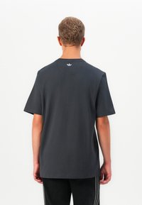 Άνδρας με την πλάτη στραμμένη, φορώντας ένα απλό σκούρο γκρι μπλουζάκι με ένα μικρό λευκό λογότυπο Adidas στην επάνω πλάτη, σε συνδυασμό με μαύρο παντελόνι.