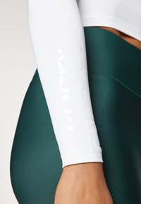 White long-sleeve top featuring a glossy "Casall" logo on the arm, paired with deep green high-waisted leggings with a smooth texture.