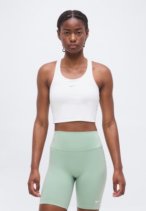 Atletyczna kobieta stoi zwrócona przodem, ubrana w biały krótki top i jasnozielone, wysokie spodenki rowerowe na jednolitym tle.