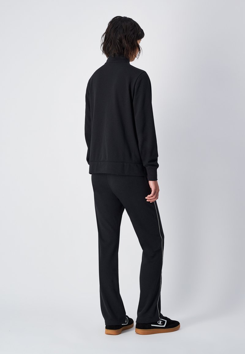 Czarny komplet swetra i spodni, z wysokim kołnierzem, jednolitym kolorem i białymi paskami po bokach. Materiał wydaje się miękki, a design jest casualowy.