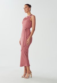 Fitted, sleeveless midi dress in soft rose with a draped neckline and a tied waist. Paired with white strappy heels. Smooth fabric texture.