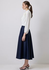 Blusa bianca con maniche lunghe e a sbuffo, abbinata a una gonna fluida blu navy, lunga fino al ginocchio. Tacchi neri con cinturini completano l'outfit.