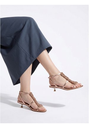 Pieds portant des sandales à talon bas couleur or rose avec des brides tressées, associées à une jupe bleu marine sur un fond uni.