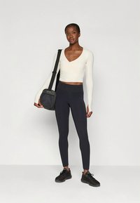 Witte top met lange mouwen en een V-hals, zwarte legging met hoge taille, en zwarte sneakers; draagt een donkere messenger bag; aansluitend model.