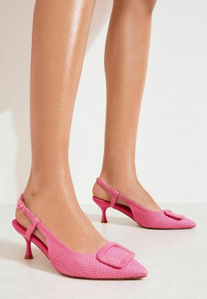 Picioare feminine purtând pantofi cu toc mic tip slingback roz texturat, cu vârf ascuțit și catarame decorative, pe un fundal alb.