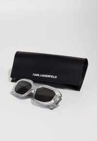 Lunettes de soleil grises avec une monture hexagonale, des détails transparents, des verres foncés et des accents en métal, accompagnées d'un étui rectangulaire noir avec un logo blanc.