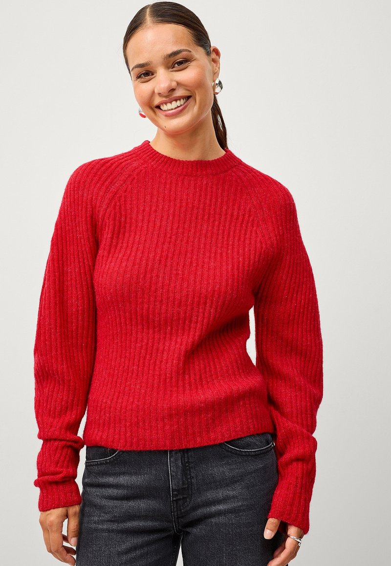 Rood geribbeld sweater met lange mouwen, een ronde hals en een relaxed fit, gecombineerd met donkere jeans. De stof lijkt textuur te hebben en is gezellig.