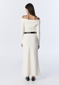 Witte gebreide jurk met een off-shoulder halslijn, lange mouwen en een zwarte riem. Gestructureerde stof en een vloerlange silhouet benadrukt.