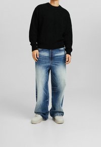 Osoba w czarnym, dzianinowym swetrze, luźnych, wyblakłych niebieskich dżinsach i brudnobiałych sneakersach stoi na jednolitym tle.