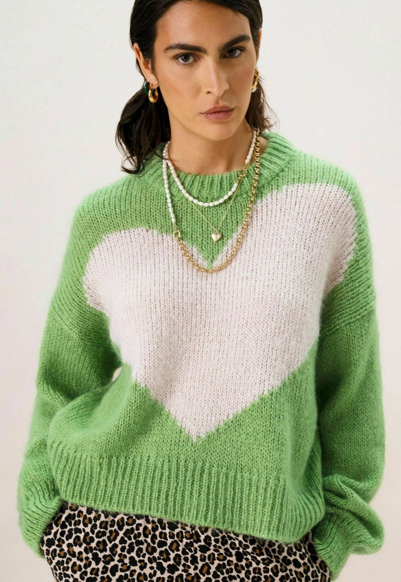 Pull en tricot vert vif avec un grand motif de cœur blanc, manches longues et bords côtelés, porté sur un bas à imprimé léopard.