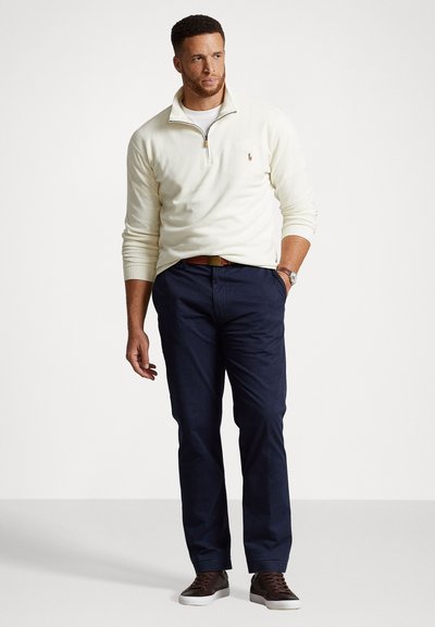 Roomwitte zip-up trui, marineblauwe broek en bruine schoenen. De trui heeft een klein logo en de broek heeft een rechte snit met riemlussen.