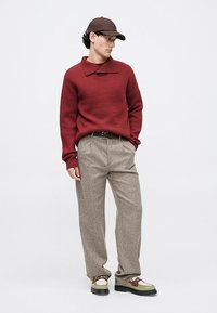 Man wearing brown cap, red knit sweater, beige trousers, and green and brown loafers standing against white background.