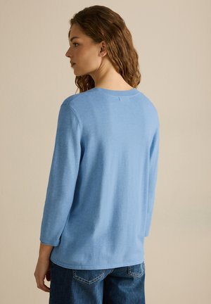Lyseblå sweater med rund halsudskæring og trekvartærmer. Blød tekstur og en afslappet pasform, parret med mørke denimjeans.