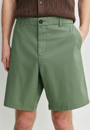 Bărbat purtând pantaloni scurți verzi de lungime medie, cu închidere frontală cu nasture și fermoar, asortați cu o cămașă tricotată maro texturată, băgată în pantaloni.