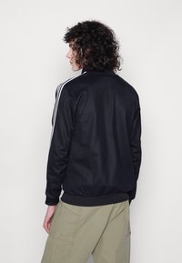 Svart jakke med høy krage, med tre hvite striper langs skuldrene. Laget av glatt stoff, med en tettsittende midje og mansjetter.