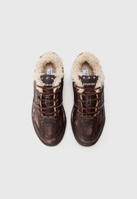 Mørkebrune lærsk sneakers med røde snørebånd og fluffy beige shearling-fôr, sett ovenfra på en hvit bakgrunn.