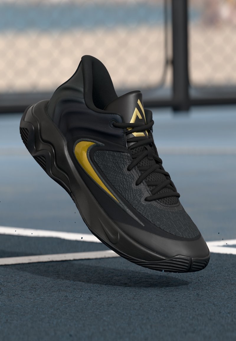 Chaussure de sport noire avec un accent doré et un logo, lacets attachés, montrée en l'air au-dessus d'une surface de terrain de sport bleue.