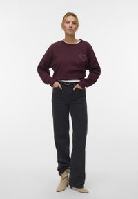 Vrouw met blond haar in een bordeauxrode sweatshirt met een hartontwerp, hoog-zittende zwarte jeans, beige laarzen en een zwarte riem die staat.