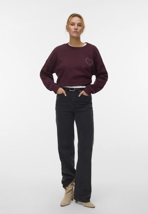 Vrouw met blond haar in een bordeauxrode sweatshirt met een hartontwerp, hoog-zittende zwarte jeans, beige laarzen en een zwarte riem die staat.