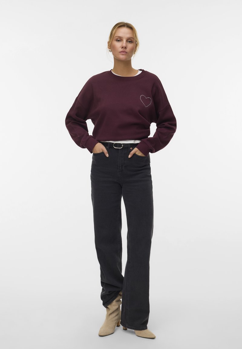 Vrouw met blond haar in een bordeauxrode sweatshirt met een hartontwerp, hoog-zittende zwarte jeans, beige laarzen en een zwarte riem die staat.