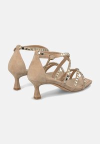Zapatos de tacón de gamuza beige con un tacón cuadrado corto, que cuentan con tiras entrelazadas y tachuelas cuadradas en tono dorado a lo largo de las tiras como detalle decorativo.