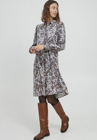 Rochie cu imprimeu floral, cu mâneci lungi, guler și tiv evazat. Prezintă un amestec de culori alb, maro și negru, asortată cu cizme maro până la genunchi.