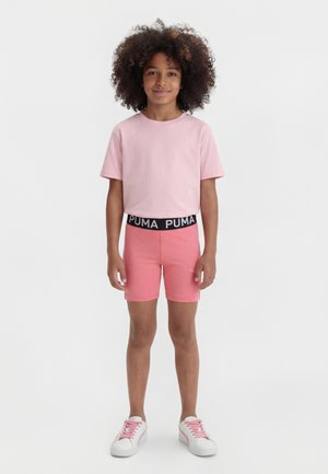 Dziecko o kręconych włosach ubrane w jasnoróżową koszulkę, różowe spodenki Puma z czarnym pasem i białe trampki z różowymi sznurowadłami.