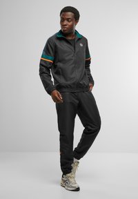 Chándal negro con acentos en verde azulado y naranja, que incluye una chaqueta con cremallera y cuello alto, y puños elásticos, combinada con pantalones ajustados.
