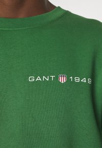 T-shirt vert col rond avec texte blanc "GANT 1949" et logo en forme de bouclier avec étoiles et rayures sur la poitrine.