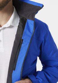 Blauwe waterdichte jas met een ritssluiting aan de voorkant, zwarte fleece voering en een kraag. Heeft een zijzak met rits en een gladde, glanzende textuur.