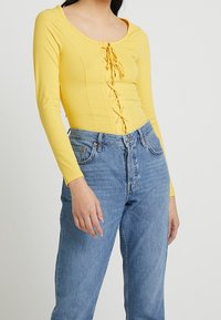 Žlutý žebrovaný bodysuit s dlouhým rukávem a výstřihem do véčka, s předním šněrováním, spojený s modrými džíny s vysokým pasem a rovnými nohavicemi.