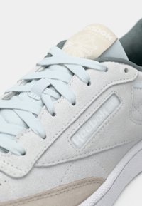 Jasnoszare zamszowe trampki z białą gumową podeszwą, z jasnoniebieskimi sznurowadłami, miękkimi beżowymi akcentami i beżowym logo na języku.