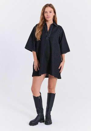 Femme portant une robe noire à fines rayures ample avec des manches larges et des bottes noires montantes jusqu'aux genoux, debout devant un fond clair uni.
