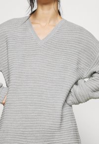 Maglione grigio chiaro a maglia con scollatura a V, maniche lunghe, texture a coste e vestibilità ampia, ideale per essere indossato a strati nelle giornate più fresche.