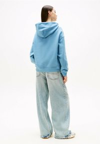 Sudadera azul clara con capucha y cordón, combinada con jeans de mezclilla claros de corte oversize. La tela parece suave, con un ajuste relajado y dobladillos sin terminar.