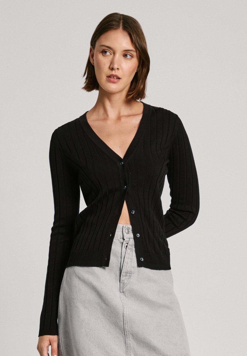 Svart ribbad cardigan med V-hals, långa ärmar och blå knappar. Bärs över en ljusfärgad kjol och har en figurnära design.