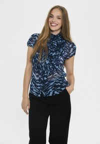 Zila rakstaina blūze ar īsiem zvīņotiem piedurkņiem, saturēta ar apkakli un tumšo un gaišo zilo toņu maisījumu, valkāta ar melniem biksēm.